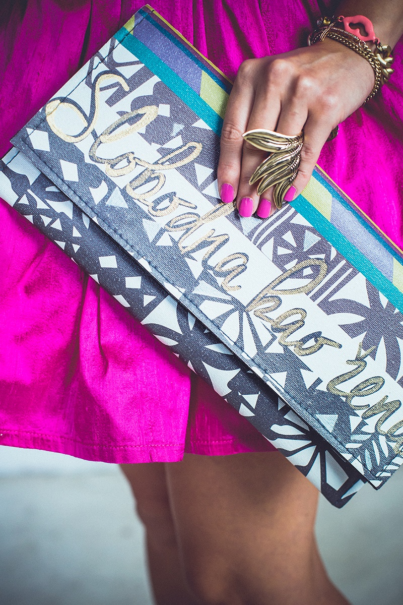 Detalj — ruka sa zlatnim prstenjem drži šarenu torbicu na magenta suknji