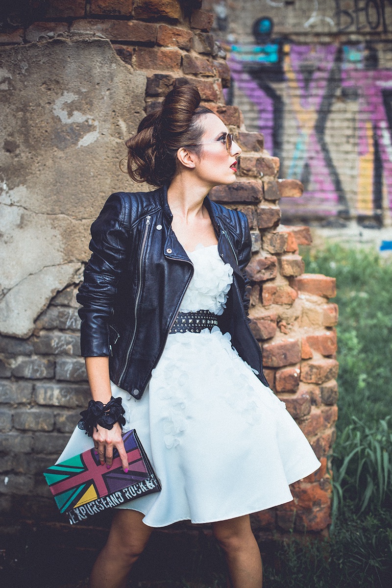Ona u beloj haljini i kožnoj jakni sa Union Jack torbom uz grafitni zid