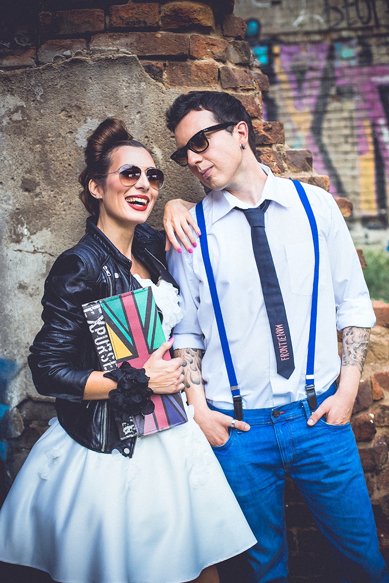 Par sa naočarima uz grafitni zid — ona sa Union Jack torbom, on sa tregerima