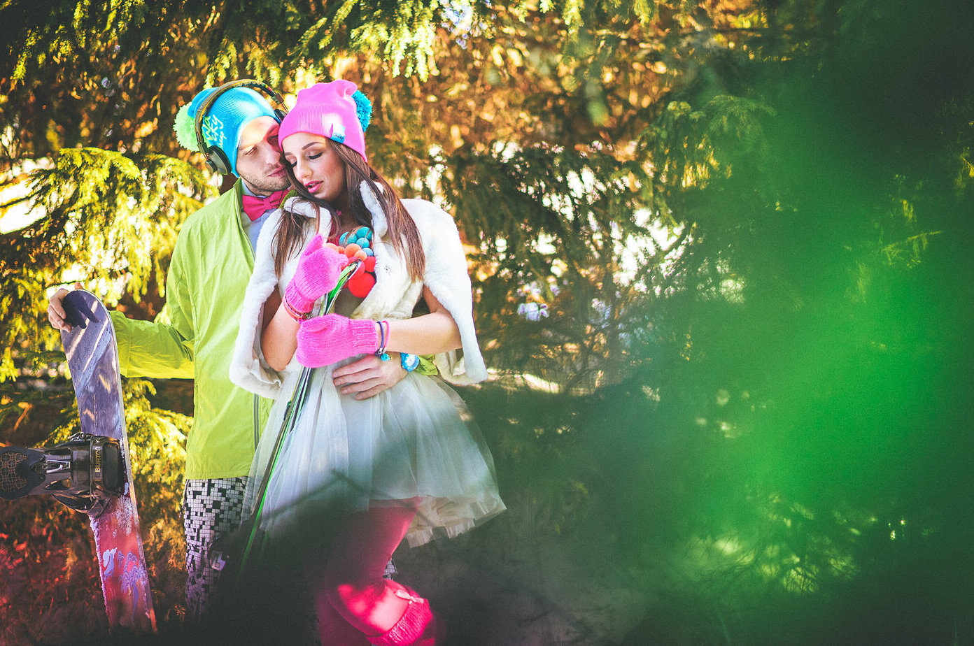 Par u zagrljaju među jelkama — on u fluo žutoj jakni sa slušalicama, ona u beloj haljini sa roze hulahopkama, lens flare u pozadini