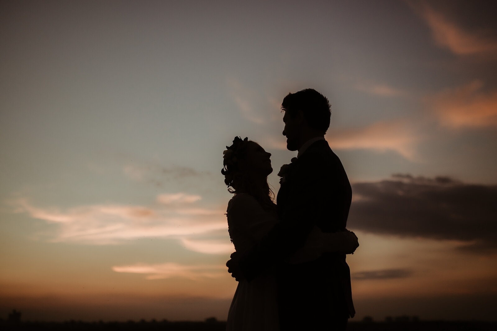 Silueta para u zagrljaju pri zalasku sunca — narandžasto-plavo nebo, cvetni venčić na glavi mlade