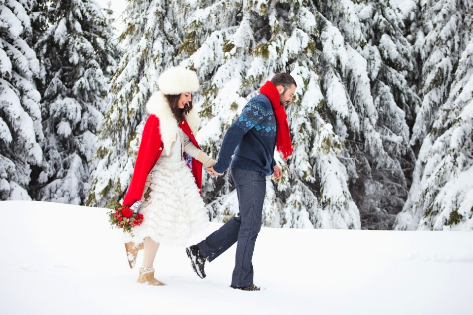 Mlada u kratkoj beloj haljini sa crvenom jaknom i mladoženja šetaju kroz duboki sneg, zasnežene jelke u pozadini